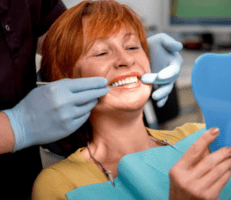 dental implant candidate smiling as she looks at her teeth in a handheld mirror